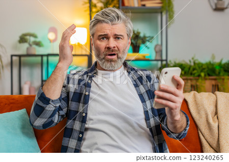Shocked sad mature man holding smartphone reading negative message trouble feels annoyed at home Shocked sad mature man holding smartphone reading negative message trouble feels annoyed at home 123240265