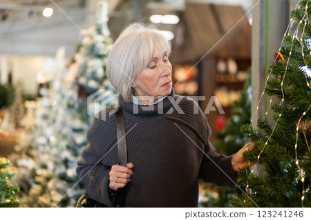 Senior woman in store examines artificial Christmas tree, examining details of product 123241246