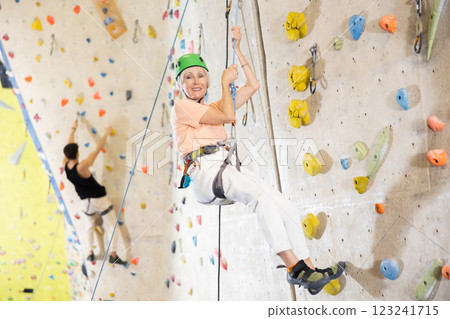elderly woman in safety helmet and with safety rope hangs near climbing wall in gym 123241715