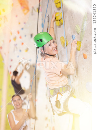 Positive senior old woman with rope training on concrete climbing rock wall to maintain healthy discipline in indoor adventure park 123241830