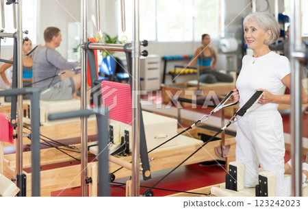 Sports club senior woman client performs exercise on reformer with cable and rope for arms Sports club senior woman client performs exercise on reformer with cable and rope for arms 123242023