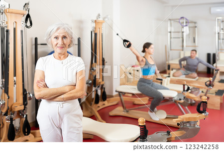 Positive senior woman coach stands in large gym against background of Pilates simulators Positive senior woman coach stands in large gym against background of Pilates simulators 123242258