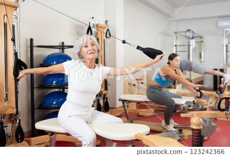 Sports club senior woman client performs exercise on reformer with cable and rope for arms Sports club senior woman client performs exercise on reformer with cable and rope for arms 123242366