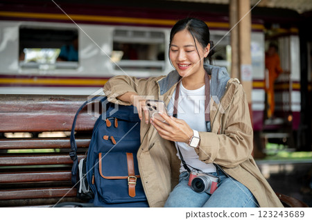 Smiling asian woman is looking through the phone while sitting on the wooden benchmark with bag. Smiling asian woman is looking through the phone while sitting on the wooden benchmark with bag. 123243689