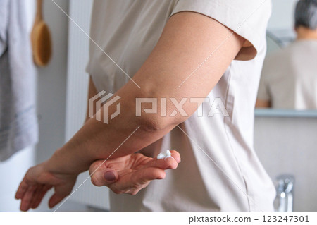 A mature woman of the pensioner age with dry skin smears moisturizing cream on her elbows. 123247301