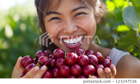 Smiling woman holding a large pile of fresh, red cherries against a backdrop of green foliage. Healthy eating and summer harvest. 123247536