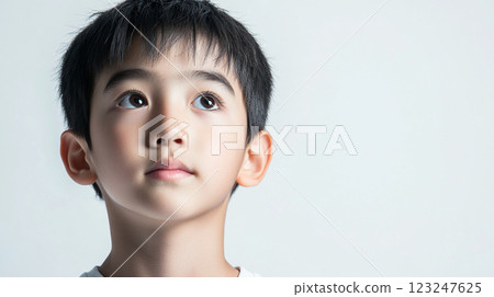 A thoughtful young boy with dark hair gazes upwards with bright, curious eyes against a plain backdrop, evoking innocence and wonder. 123247625