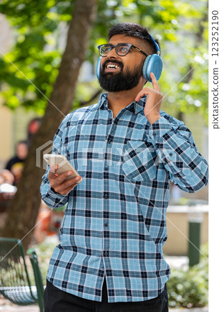 Happy Indian man dancing in wireless headphones listening favorite energetic music on city street Happy Indian man dancing in wireless headphones listening favorite energetic music on city street 123252190