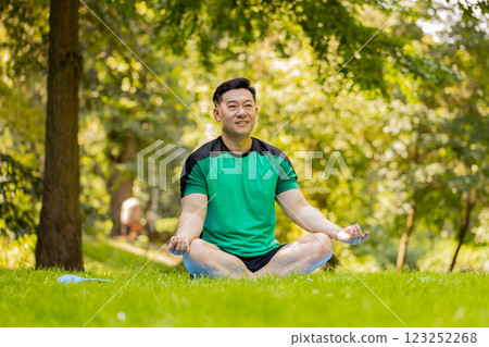 Young Asian man sitting on mat in lotus position, relaxing, practicing yoga in park on summer day 123252268