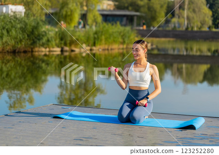 Sporty young woman exercising with dumbbells kneeling on yoga mat near pond in park on sunny day Sporty young woman exercising with dumbbells kneeling on yoga mat near pond in park on sunny day 123252280