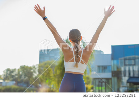 Rear view of young woman runner raising arms celebrating success after jogging in urban city park Rear view of young woman runner raising arms celebrating success after jogging in urban city park 123252281