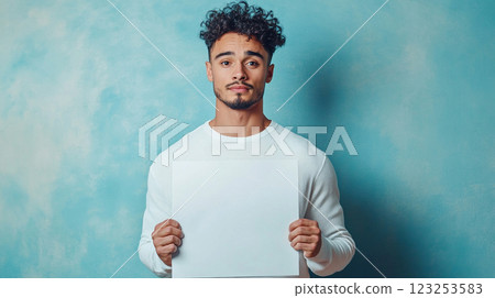 Person holding blank white paper against textured turquoise background looking at the camera with a serious expression. 123253583