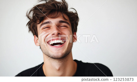 A joyful young man with curly brown hair and a black t-shirt is laughing with his eyes closed against a plain background. 123253873