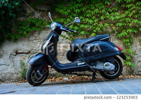 Black scooter parked on cobblestone street against old brick wall covered in green ivy. Vintage urban transportation 123253903