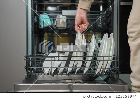 Woman unloading clean white plates from the bottom rack of dishwasher in kitchen. Person performing everyday household chores, using modern home appliance Woman unloading clean white plates from the bottom rack of dishwasher in kitchen. Person performing everyday household chores, using modern home appliance 123253974