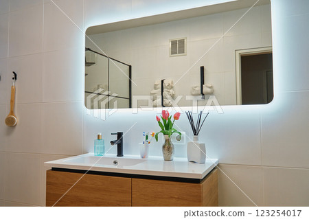 Stylish bathroom with wooden sink cabinet, black faucet and decorative elements. Contemporary interior design of bathroom with illuminated large LED mirror in modern apartment 123254017