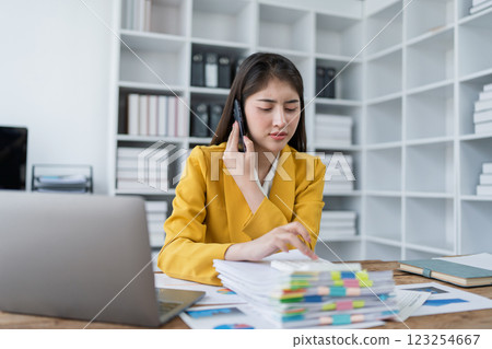 Businesswoman in a yellow suit making a phone call while organizing documents in a modern office workspace Businesswoman in a yellow suit making a phone call while organizing documents in a modern office workspace 123254667