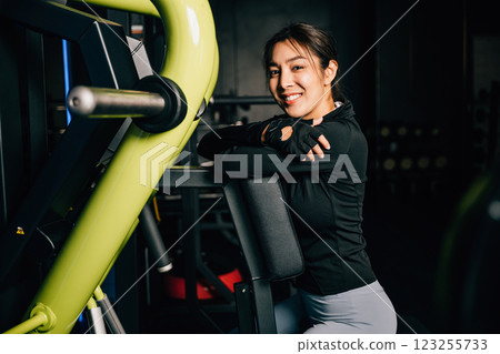 Portrait of beautiful Asian young woman performing squats on a gym machine, female working out with rowing machine at crossfit fitness, healthy lifestyle concept Portrait of beautiful Asian young woman performing squats on a gym machine, female working out with rowing machine at crossfit fitness, healthy lifestyle concept 123255733