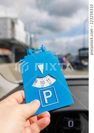 Driver hand hold blue parking disc show arrival time near large shopping complex mall. Parking time restrictions in busy parking lots. Limited time duration park place zone Driver hand hold blue parking disc show arrival time near large shopping complex mall. Parking time restrictions in busy parking lots. Limited time duration park place zone 123256310