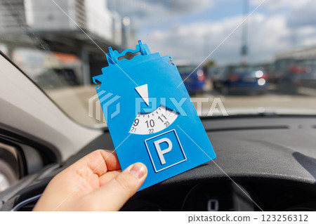 Driver hand hold blue parking disc show arrival time near large shopping complex mall. Parking time restrictions in busy parking lots. Limited time duration park place zone Driver hand hold blue parking disc show arrival time near large shopping complex mall. Parking time restrictions in busy parking lots. Limited time duration park place zone 123256312