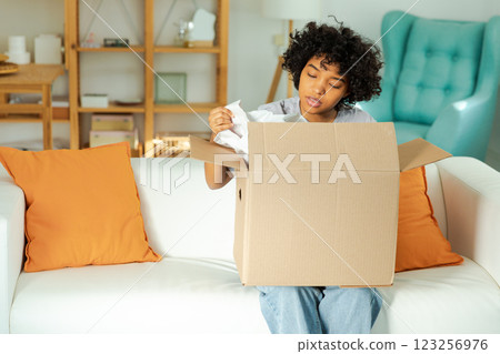 African girl unpacking delivery looking in box. Happy woman opening carton box. Female getting parcel looking at delivered goods items. Satisfied client positive feedback quick express deliver 123256976