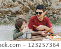 Close-up view of father hugs his school boy son on a family picnic. Child kid and his dad taking a rest and enjoying a picnic while hiking. Boy smiles and bites the bread donut on a picnic 123257364