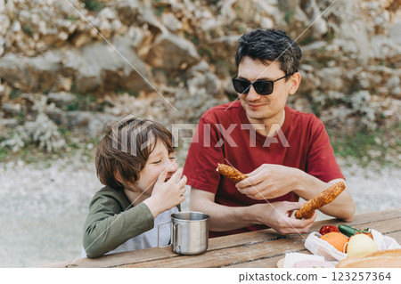 Close-up view of father hugs his school boy son on a family picnic. Child kid and his dad taking a rest and enjoying a picnic while hiking. Boy smiles and bites the bread donut on a picnic 123257364