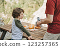 Father dad pours hot coffee tea from thermos into the mug on a family picnic in the mountains. Child school boy kid is watching his dad filling the cup with hot beverage 123257365