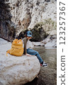 School boy with yellow backpack sits on a riverside rock in the canyon with mountain cliffs in the background. Kid child taking a rest on a boulder near mountain river 123257367