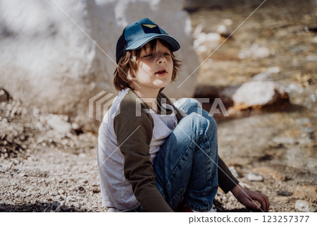 Caucasian school boy sits on a riverside rock in the canyon with mountain cliffs in the background. Kid child taking a rest on a boulder near mountain river bank Caucasian school boy sits on a riverside rock in the canyon with mountain cliffs in the background. Kid child taking a rest on a boulder near mountain river bank 123257377