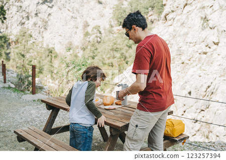 Father dad pours hot coffee tea from thermos into the mug on a family picnic in the mountains. Child school boy kid is watching his dad filling the cup with hot beverage 123257394