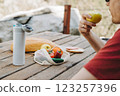 Adult man in sunglasses eating bread on a family picnic with coffee, bread, and vegetables on the wooden table 123257396