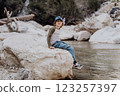 Caucasian school boy sits on a riverside rock in the canyon with mountain cliffs in the background. Kid child taking a rest on a boulder near mountain river bank 123257397