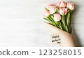 A bouquet of pink tulips wrapped in a light craft paper sits on a white wooden surface. A tag attached reads 'Happy Mother's Day,' celebrating the occasion with elegance. 123258015