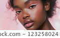 A close-up portrait of a young Black woman with beautiful skin and stylish makeup. The makeup features pastel colors, bold eyeliner, and glossy lips. The background is soft pink, enhancing her 123258024