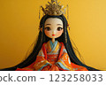 A beautiful traditional doll wearing a colorful robe and an ornate crown. The doll features long, black hair and a calm expression. The background is a solid yellow color. 123258033