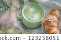 A vibrant green matcha latte rests in a cup adorned with floral latte art. Next to it lies a fresh, flaky croissant. The scene features soft pastel colors and natural light. 123258051