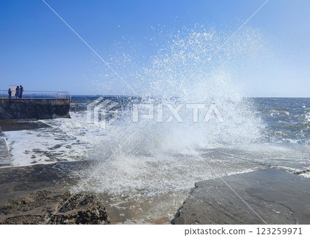 Storm at sea on a sunny summer day. Big sea waves with white foam crashing at concrete old pier on a sunny day. Lots of splashes from big waves crashing against the cement pier and clean blue sky. Storm at sea on a sunny summer day. Big sea waves with white foam crashing at concrete old pier on a sunny day. Lots of splashes from big waves crashing against the cement pier and clean blue sky. 123259971