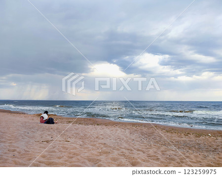 Woman sitting on sandy beach on seashore near sea with waves during rain. Dark storm clouds, rain and sea surface at sunset. Nature. Natural background. Stormy weather. Storm at sea. 123259975