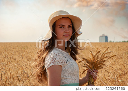 Young woman in sun hat holding wheat in golden field under blue sky. Harvesting concept 123261360