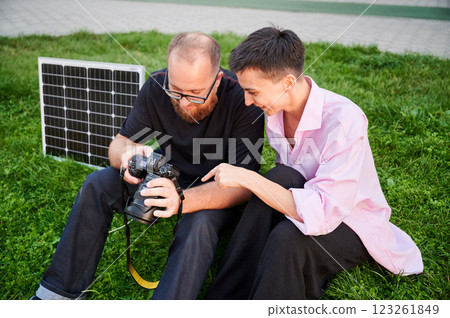 Couple sits on grass, closely examining photos on camera, with solar panel nearby. Integration of renewable energy in photography. Sustainable charging solutions for photography equipment. Couple sits on grass, closely examining photos on camera, with solar panel nearby. Integration of renewable energy in photography. Sustainable charging solutions for photography equipment. 123261849