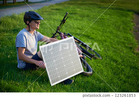 Man cyclist using solar panel for charging electric mountain bike outdoors. Guy wearing helmet and sunglasses sits on grass in park, enjoying sunny evening. Man cyclist using solar panel for charging electric mountain bike outdoors. Guy wearing helmet and sunglasses sits on grass in park, enjoying sunny evening. 123261853