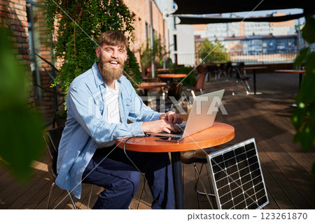 Man works on laptop and smartphone at outdoor table, with glass of iced coffee. Solar panel nearby, emphasizing sustainable, modern eco-friendly workspace that combines technology and sustainability. Man works on laptop and smartphone at outdoor table, with glass of iced coffee. Solar panel nearby, emphasizing sustainable, modern eco-friendly workspace that combines technology and sustainability. 123261870