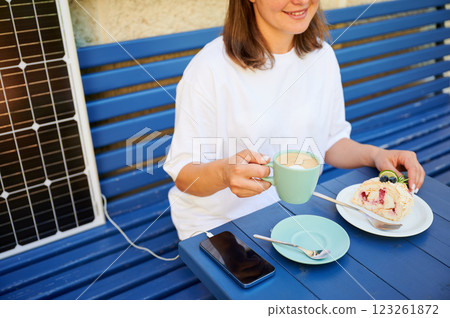 Girl drinking coffee while phone charging from solar panel. Cropped view of happy woman sitting next to portable solar battery charger. Integration of sustainable renewable energy into everyday life. Girl drinking coffee while phone charging from solar panel. Cropped view of happy woman sitting next to portable solar battery charger. Integration of sustainable renewable energy into everyday life. 123261872