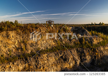 A rocky cliff with sparse trees and dry bushes under warm sunlight. At the top, an abandoned industrial structure stands among autumn foliage, creating a contrast between nature and human-made ruins. A rocky cliff with sparse trees and dry bushes under warm sunlight. At the top, an abandoned industrial structure stands among autumn foliage, creating a contrast between nature and human-made ruins. 123263385