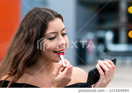 Young Woman Applying Lipstick While Checking Reflection on Smartphone Outdoors 123264084