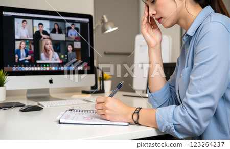 Professional Asian woman in home office writing notes while participating in virtual meeting, multiple monitors displaying video calls, modern workspace 123264237
