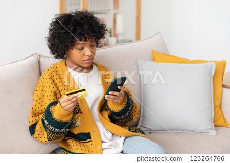 African american woman shopping online holding smartphone paying with gold credit card. Girl sitting at home buying on Internet enter credit card details. Online shopping ecommerce delivery service African american woman shopping online holding smartphone paying with gold credit card. Girl sitting at home buying on Internet enter credit card details. Online shopping ecommerce delivery service 123264766