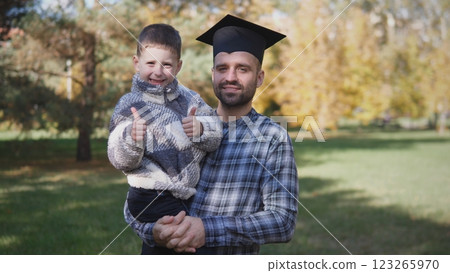 A joyous graduate proudly holds his beloved son, celebrating their success together amidst the beautiful autumn scenery A joyous graduate proudly holds his beloved son, celebrating their success together amidst the beautiful autumn scenery 123265970