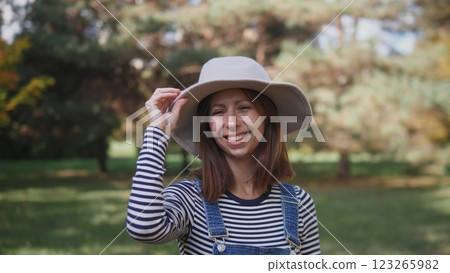 A joyful woman, wearing a fashionable hat and a striped shirt, poses happily in a serene outdoor setting 123265982